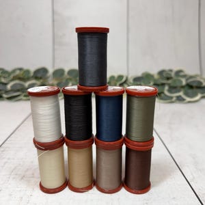 May include: A collection of spools of thread in various colors, including white, black, navy, and olive green. The spools have red tops and are stacked on a white wooden surface. The thread is likely for sewing or crafting projects.