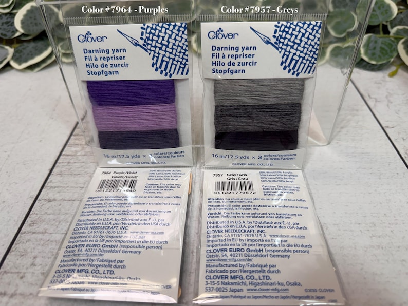 Op de afbeelding: Twee verpakkingen Clover stopgaren in doorzichtig plastic. E&eacute;n verpakking is gelabeld "Color #7964 - Purples" en bevat paars garen. De andere is gelabeld "Color #7957 - Greys" en bevat grijs garen. Elke verpakking bevat 16 m garen.