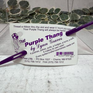 May include: A purple sewing tool called "Purple Thang" by Lynn Graves, with a slot for threading ribbon. The tool is displayed with its product information card, which includes the text "Thread a ribbon thru the slot and wear it 'round your neck."