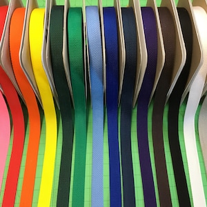 Puede incluir: Una exhibición de rollos de cincha coloridos en varios tonos, incluyendo rosa, rojo, naranja, amarillo, verde, azul y marrón. La cincha está cuidadosamente dispuesta en soportes de cartón individuales, mostrando un espectro de colores para manualidades.