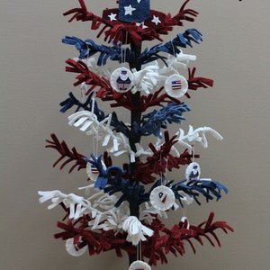 May include: A red, white and blue felt Christmas tree with a star-shaped topper and decorations. The tree is decorated with felt ornaments featuring stars and the American flag. The tree is on a white felt base with red and blue stars.