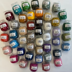 May include: A collection of colorful embroidery thread spools in various shades, including green, blue, yellow, and red. Each spool is individually packaged and labeled with a product code. The threads are neatly arranged, creating a visually appealing display.