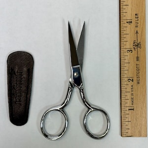 May include: A pair of silver-colored, small, sharp scissors with a leather sheath. The scissors are about 3 inches long and are next to a ruler.