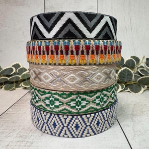 Puede incluir: Una pila de seis cintas decorativas tejidas con varios patrones y colores. La cinta superior presenta un diseño en zigzag negro, gris y blanco. Otras cintas muestran patrones geométricos y florales en tonos de azul, verde, beige, naranja y rojo.