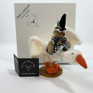 Peut inclure: Une oie blanche aux pieds orange portant une robe à carreaux noirs et blancs, un chapeau de sorcière et chevauchant l'oie. L'oie est sur une base en bois. L'oie a des ailes blanches. L'oie est une pièce de collection du D.C.A.D. Collectors Club.