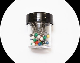 Peut inclure: Un récipient en plastique transparent rempli d'épingles de couture. Les épingles ont des tiges argentées et des têtes rondes colorées de différentes couleurs, dont le rouge, le vert, le jaune et le bleu. Le récipient a un couvercle noir.