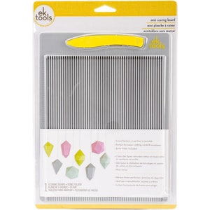 May include: A mini scoring board with a yellow bone folder. The board has vertical score lines and a ruler along the side. The packaging includes text in English and Spanish, highlighting its use for paper crafting, cards, and envelopes.