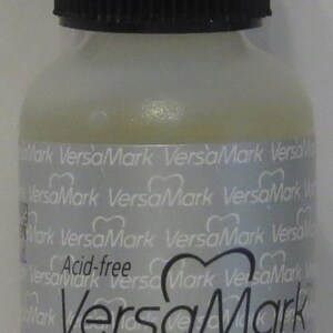 May include: A clear plastic bottle with a black screw-top lid. The bottle is labelled "VersaMark" and "Watermark ink refill." The label also states "Acid-free" and "0.5 fl.oz (15ml)".