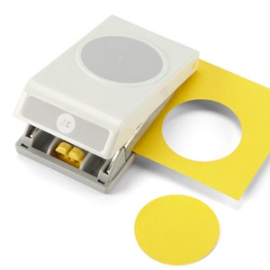 May include: A white and gray circle paper punch with a yellow circle cut-out and a yellow square paper with a circle hole. The punch has the number "2" printed on it. The cut-out circle is approximately 2 inches in diameter.