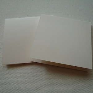 May include: Two blank white greeting cards, folded in half, are stacked on top of each other. The cards are on a textured white surface.