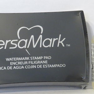 May include: A black ink pad with the text "VersaMark" and "Watermark Stamp Pad" in white. The ink pad is next to a clear bottle of VersaMark Watermark Ink Refills, which is labelled "Acid-free" and has a capacity of 0.5 fl oz (15 ml).