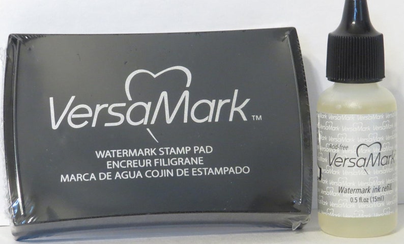 May include: A black ink pad with the text "VersaMark" and "Watermark Stamp Pad" in white. The ink pad is next to a clear bottle of VersaMark Watermark Ink Refills, which is labelled "Acid-free" and has a capacity of 0.5 fl oz (15 ml).