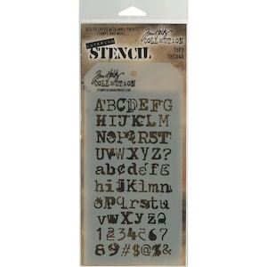 Puede incluir: Una hoja de plantilla con fondo dorado y letras negras. La plantilla incluye el alfabeto en mayúsculas y minúsculas, números y símbolos. El texto "Jim Holtz Collection" y "Layering Stencil" está impreso en la hoja.