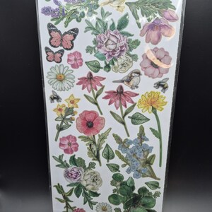 May include: A sheet of floral and fauna stickers in a clear plastic package. The stickers feature watercolor illustrations of pink and purple flowers, butterflies, bees, and birds. The stickers are on a white background.