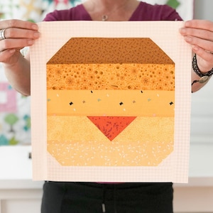 Peut inclure: Un carré de tissu rose avec un motif de patchwork représentant un gâteau à étages. Le gâteau est fait de tissu brun, jaune et orange avec un triangle rouge au centre.