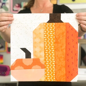 May include: A patchwork pumpkin quilt block with orange, brown, and white fabric. The pumpkin is made up of different patterns and textures, including circles, hexagons, and stripes.