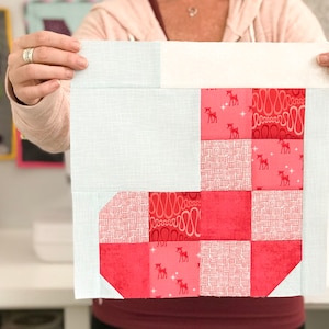 May include: A light blue fabric square with a white border. The center of the square is a patchwork of red fabric squares with different patterns, including a white and red geometric pattern, a red pattern with white reindeer, and a red pattern with white swirls.