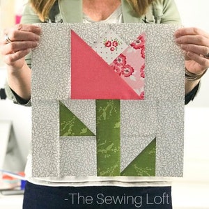 May include: A square quilt block featuring a tulip design. The tulip has a pink flower with floral patterned accents and green stem and leaves. The background is a light gray fabric with a leaf pattern. The text "-The Sewing Loft" is at the bottom.