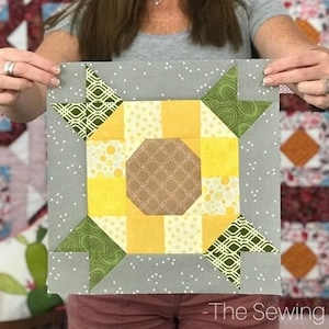 Pode incluir: Um bloco de colcha quadrado com um desenho de girassol. O centro é castanho, rodeado por quadrados amarelos e triângulos verdes. O bloco está sobre um fundo cinzento com bolinhas brancas. O logotipo The Sewing Loft está na parte inferior.