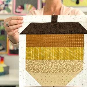 Op de afbeelding: Een bruine, gele en witte patchwork eikelblok voor een quilt. De blok is gemaakt van verschillende stoffen met verschillende patronen, waaronder stippen, strepen en bloemenpatronen. De blok is omgeven door een witte achtergrond met kleine witte stippen.