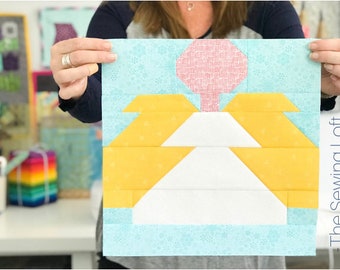 May include: A quilt block with a white, yellow, and pink design. The block is made up of squares and triangles. The block is on a blue background with a white pattern. The text "The Sewing Loft" is on the right side of the image.