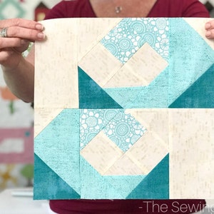 May include: Two quilt blocks with a geometric design. Each block features a central square with a light blue and white floral pattern, surrounded by teal and cream-colored fabric. The blocks are held up by a person.