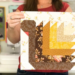 May include: A square quilt block featuring a turkey design. The turkey is constructed from various fabric patterns, including floral, striped, and solid colors in shades of brown, yellow, and cream. The background is a cream color with a floral pattern.