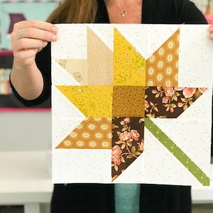 May include: A patchwork quilt block featuring a star design. The star is composed of various patterned fabrics in shades of yellow, brown, and floral prints, set against a white background with small, scattered dots. A green stripe adds a contrasting element.