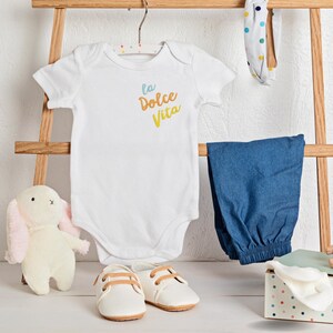 May include: A white baby onesie with the text "la Dolce Vita" in blue and yellow, paired with blue pants, white shoes, and a stuffed bunny. A polka-dot headband and a small gift box are also present.