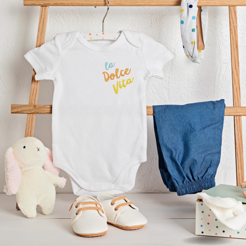 May include: A white baby onesie with the text "la Dolce Vita" in blue and yellow, paired with blue pants, white shoes, and a stuffed bunny. A polka-dot headband and a small gift box are also present.