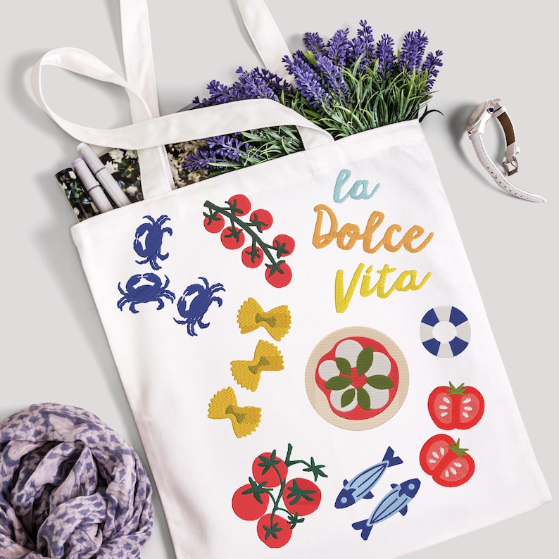 May include: White tote bag with a variety of colorful embroidered Italian food and beach-themed designs. The bag features images of crabs, tomatoes, pasta, and the phrase "La Dolce Vita". The bag is filled with lavender and pens.