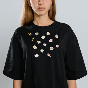 May include: Black oversized t-shirt with small, colorful embroidered illustrations of teacups, teapots, cakes, and flowers. The shirt has short sleeves and a crew neck, offering a casual and comfortable style.