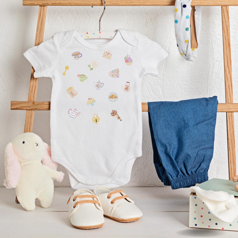 May include: A white baby bodysuit with colorful food and tea-themed illustrations hangs on a wooden rack, alongside blue pants, white shoes, a bunny plush, and a polka-dot headband. The baby clothes are arranged on a white surface.