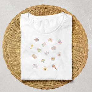 May include: White t-shirt with various embroidered pastel-colored illustrations of teacups, teapots, pastries, and flowers. The shirt is folded on a round, woven wicker surface.