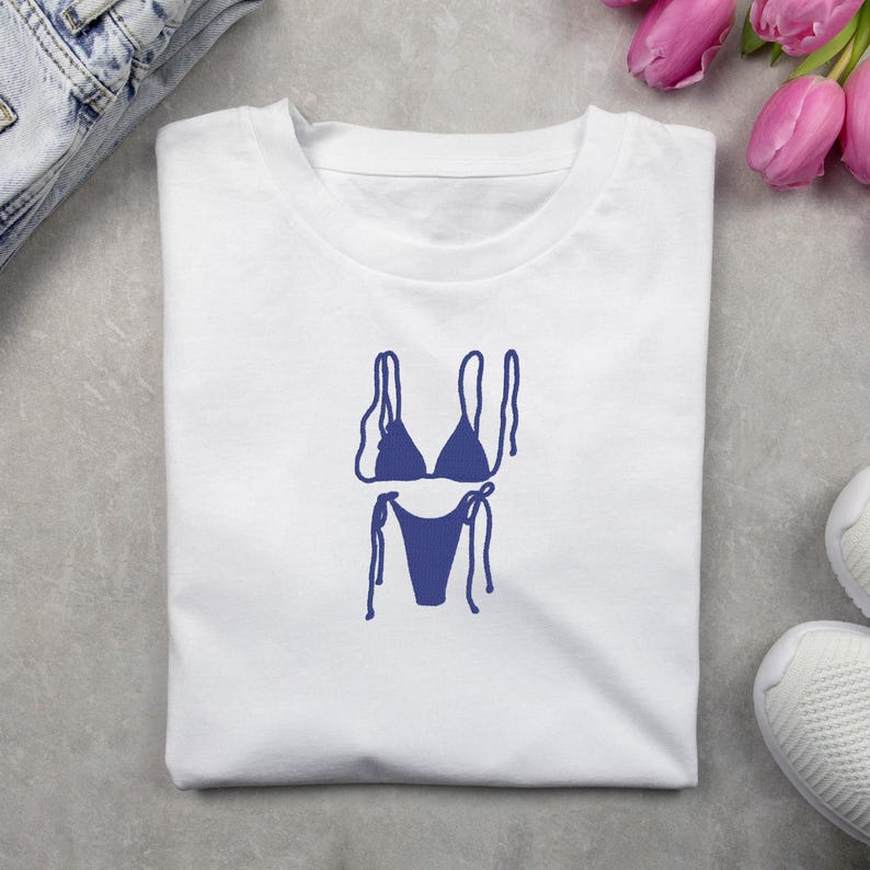 May include: White t-shirt with a navy blue bikini design. The graphic shows a two-piece swimsuit with tie details. The shirt is folded, with jeans and a trainer visible in the background.