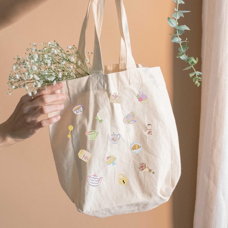 May include: Cream-colored tote bag adorned with colorful embroidered food and drink motifs, including teacups, teapots, and pastries. The bag has long handles and is filled with white flowers. A charming and practical accessory.