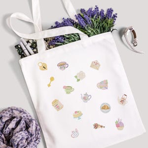 May include: A white tote bag with various embroidered tea party-themed designs, including teacups, teapots, and pastries. The bag is filled with lavender and pens. A watch and a scarf are also visible.