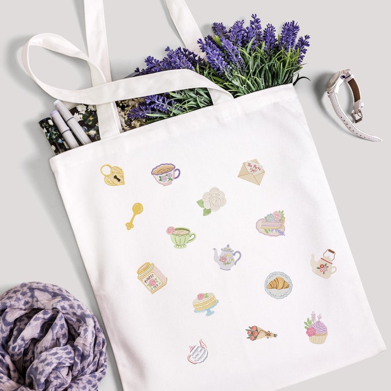 May include: A white tote bag with various embroidered tea party-themed designs, including teacups, teapots, and pastries. The bag is filled with lavender and pens. A watch and a scarf are also visible.