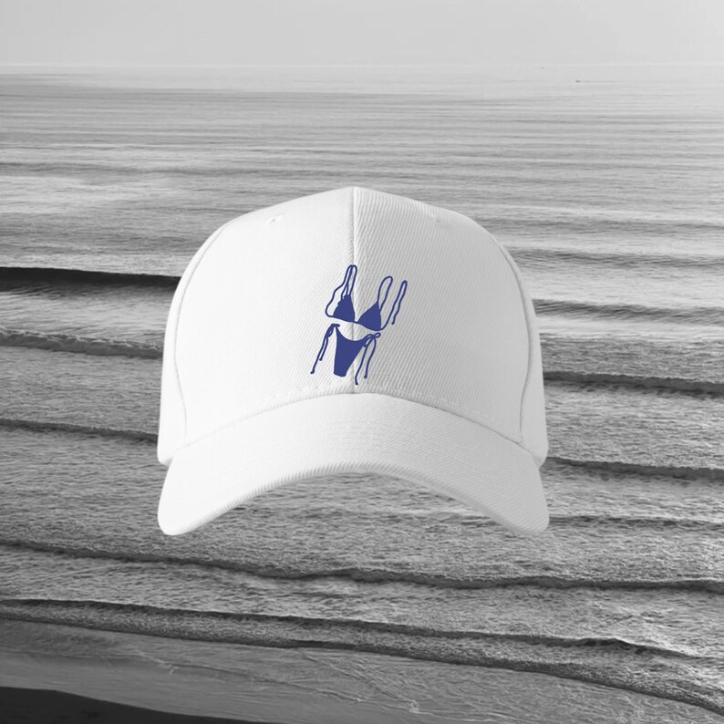 May include: White baseball cap with a blue outline of a bikini. The cap has a curved peak and a structured crown. The background is a grayscale image of the sea.
