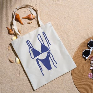 May include: A cream tote bag featuring a navy blue bikini design, seashells, a straw hat, and sunglasses on sand. Ideal for beach trips, the bag's design is simple and stylish.
