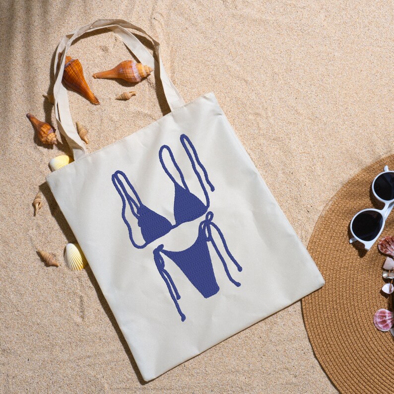 May include: A cream tote bag featuring a navy blue bikini design, seashells, a straw hat, and sunglasses on sand. Ideal for beach trips, the bag's design is simple and stylish.