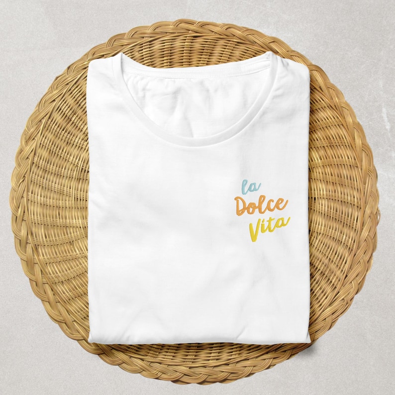 May include: A white t-shirt with the phrase "la Dolce Vita" embroidered in blue, orange, and yellow. The shirt is folded and placed on a round, woven wicker surface.