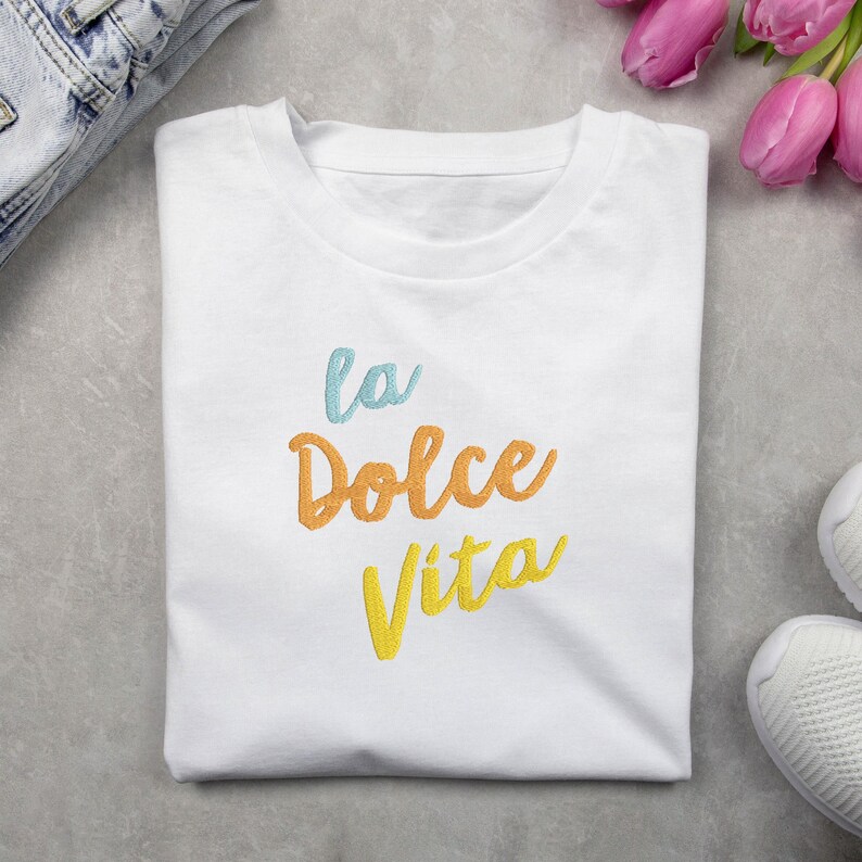 May include: White t-shirt with the embroidered phrase "La Dolce Vita" in blue, orange, and yellow script. The shirt is folded on a gray surface, with a pair of jeans and a bouquet of pink tulips in the background.