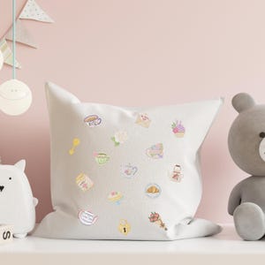 May include: A white square pillow decorated with embroidered pastel-colored illustrations of teacups, teapots, cakes, and flowers. A teddy bear and other decorative items are in the background. The pillow is on a white surface.