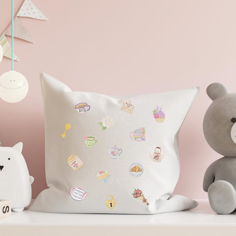 May include: A white square pillow decorated with embroidered pastel-colored illustrations of teacups, teapots, cakes, and flowers. A teddy bear and other decorative items are in the background. The pillow is on a white surface.