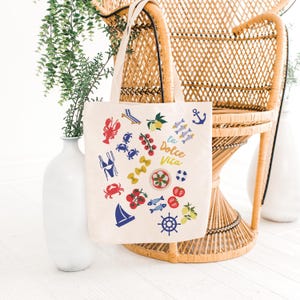 May include: Cream-colored tote bag with colorful Italian-themed illustrations. The bag features images of a lobster, crabs, tomatoes, pasta, fish, lemons, and a sailboat. The words "la Dolce Vita" are printed on the bag.