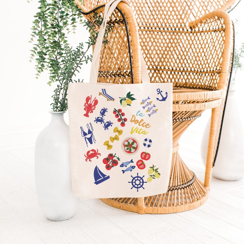 May include: Cream-colored tote bag with colorful Italian-themed illustrations. The bag features images of a lobster, crabs, tomatoes, pasta, fish, lemons, and a sailboat. The words "la Dolce Vita" are printed on the bag.