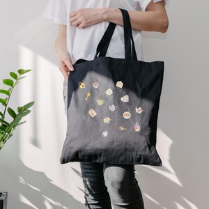 May include: Black tote bag with a collection of colorful, whimsical food and tea-themed illustrations. The bag has two black handles and is held by a person wearing a white shirt and gray jeans. The illustrations include teacups, cakes, and teapots.