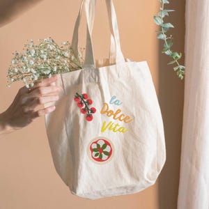 May include: Cream-colored tote bag with the phrase "la Dolce Vita" in blue, orange, and yellow. The bag features illustrations of tomatoes and a pizza-like design. The bag is filled with white flowers.