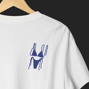May include: White t-shirt with a navy blue bikini graphic on the back. The bikini design is a simple line drawing, showing the top and bottom with tie details. The shirt is hanging on a wooden hanger against a dark background.